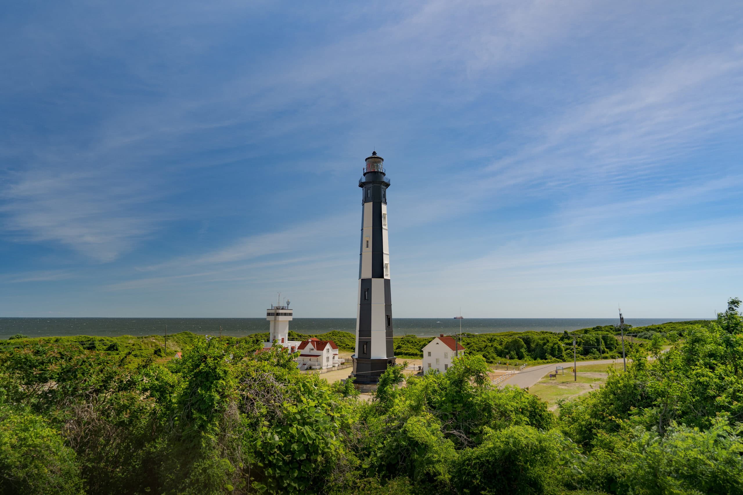 A coastal landscape featuring a tall black and white lighthouse surrounded by greenery, with a view of the ocean and a clear blue sky.