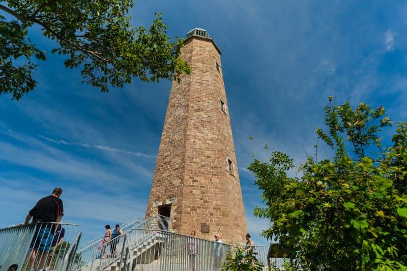 Tall stone lighthouse with steps leading up to it, surrounded by greenery and people, set against a blue sky with clouds.