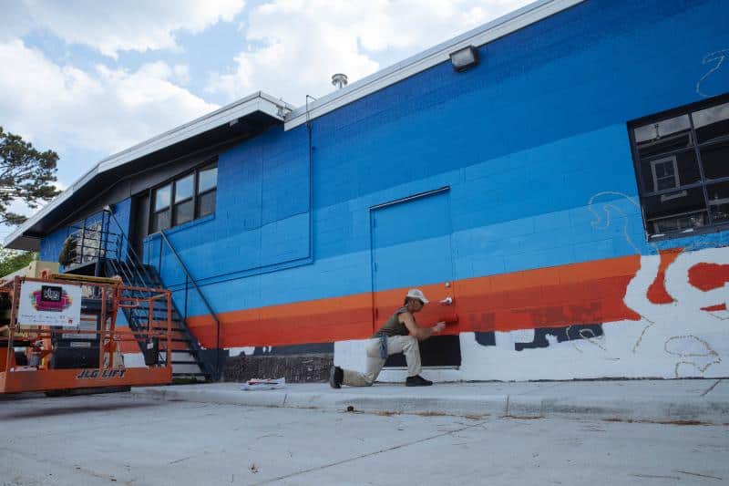 An artist paints a colorful mural on a building using a ladder platform. The artwork includes blue, orange, and red stripes.