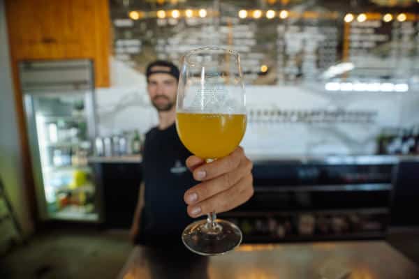 Person holding a glass of yellow beer in a bar with a chalkboard menu in the background.