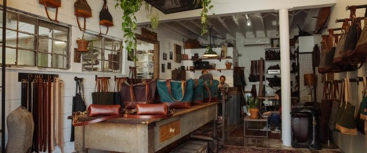 Leather goods store interior with various bags displayed on tables and shelves. Hanging plants and rustic decor present, creating a warm and inviting atmosphere.