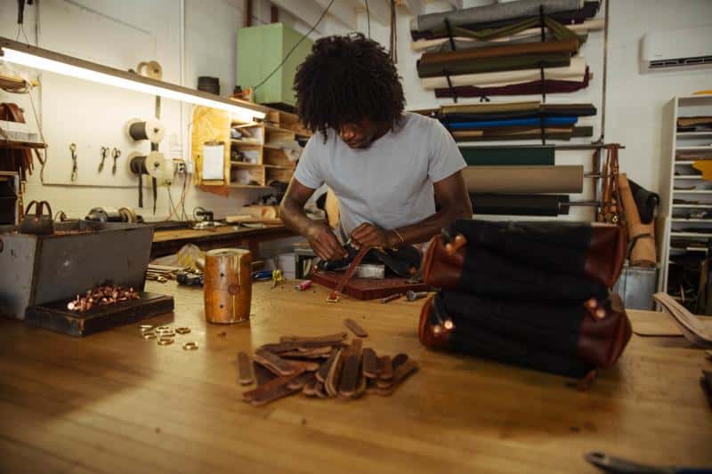 Person working on leather craft at a wooden table in a workshop, surrounded by tools, cut leather pieces, and completed bags.