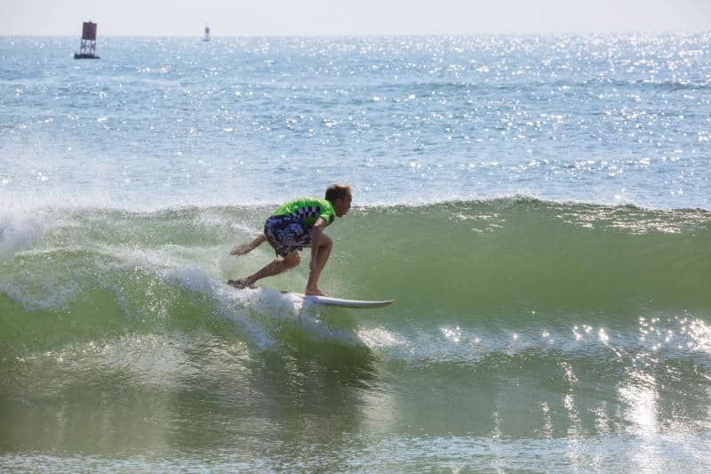A surfer wearing a green shirt rides a wave on a sunny day. The ocean is calm with a distant buoy visible on the horizon.