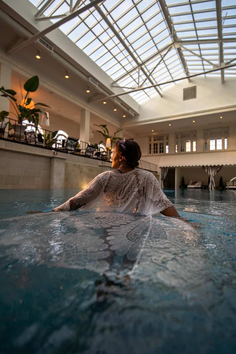 Person wearing a lace garment stands waist-deep in an indoor pool, gazing up. The room features a glass ceiling, potted plants, and arches.