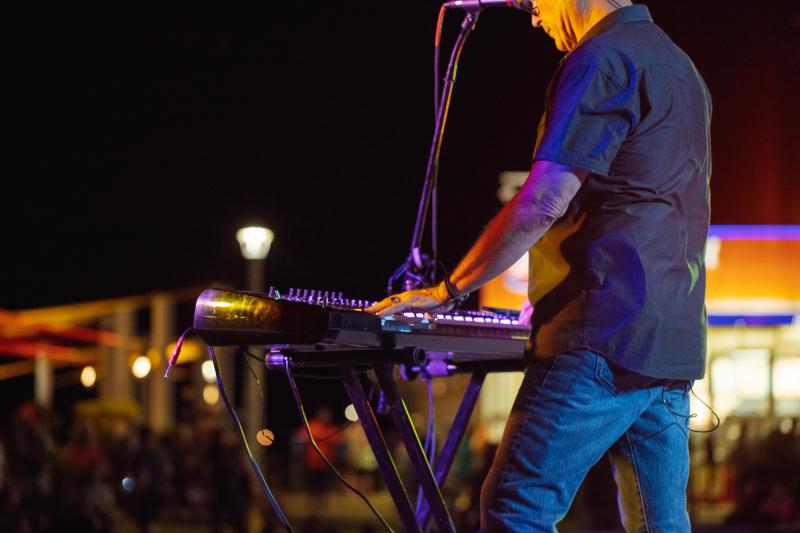 A musician plays a keyboard on stage at night, with colorful lights in the background.