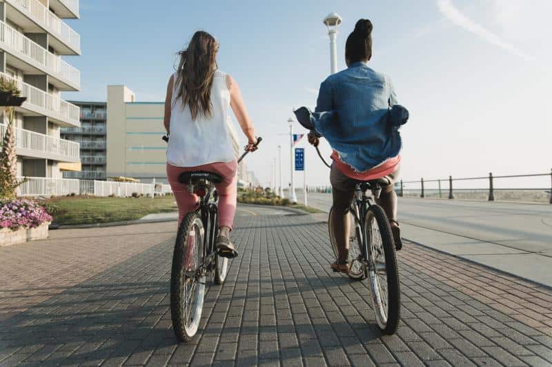 Two people riding bicycles on a paved path beside a beach, beachfront buildings on the left.