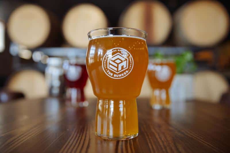A glass of amber beer with "The Bunker" logo on a wooden table, surrounded by blurred beer barrels in the background.