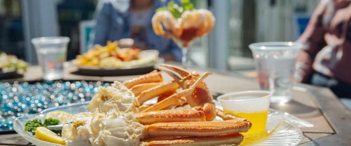 A plate of crab legs with butter and lemon is on the table. In the background are two people, a cup of shrimp cocktail, and a dish of unidentified food.