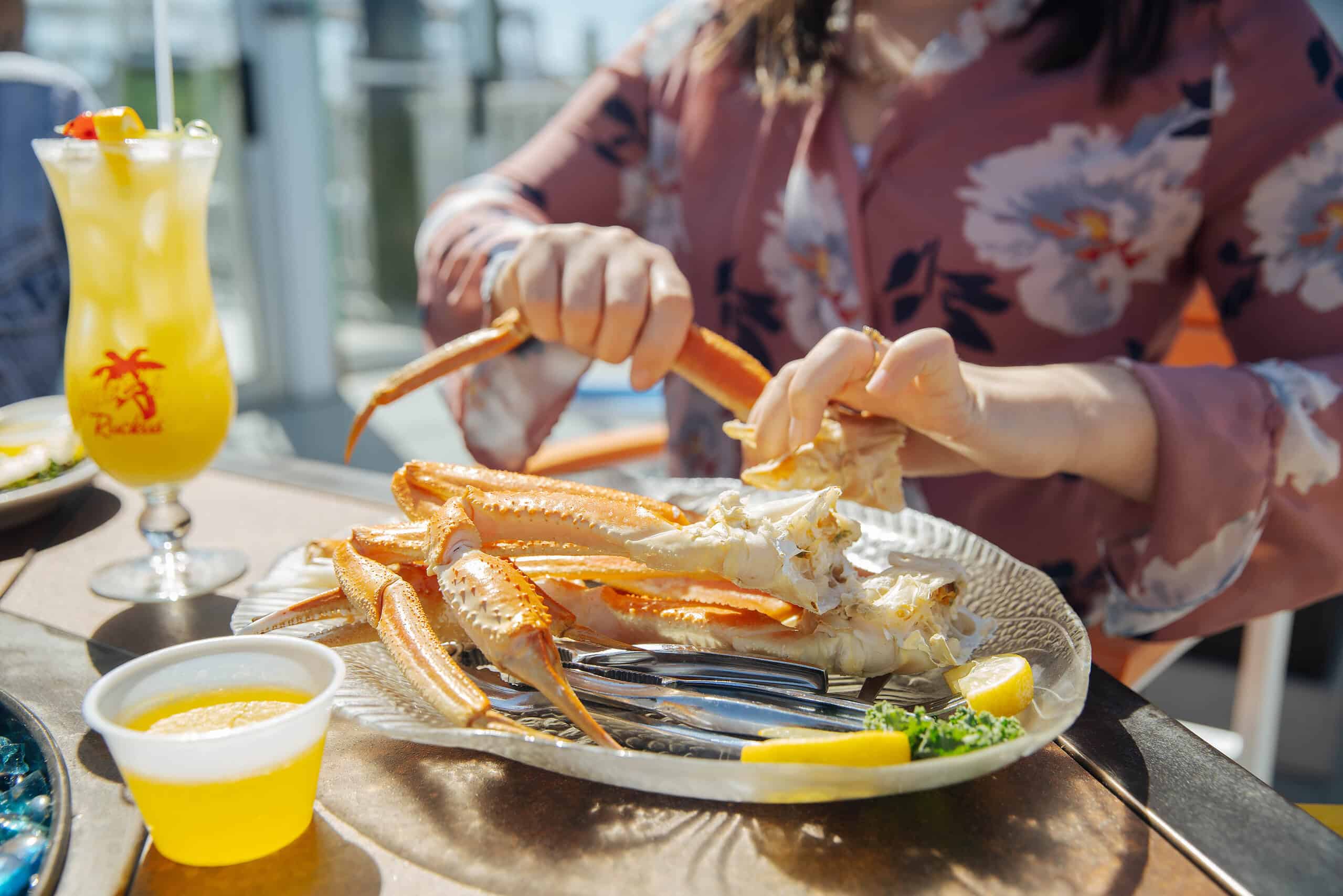 At Rudee's Restaurant & Cabana Bar a person cracking crab legs on a plate with lemon wedges and parsley. Beside the plate is a drink in a tall glass and a small cup of butter.