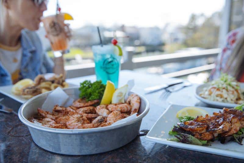 A table with grilled shrimp in a metal bowl, garnished with lemon and greens, beside a blue cocktail and other dishes. Person in background drinking from a tall glass with a straw.