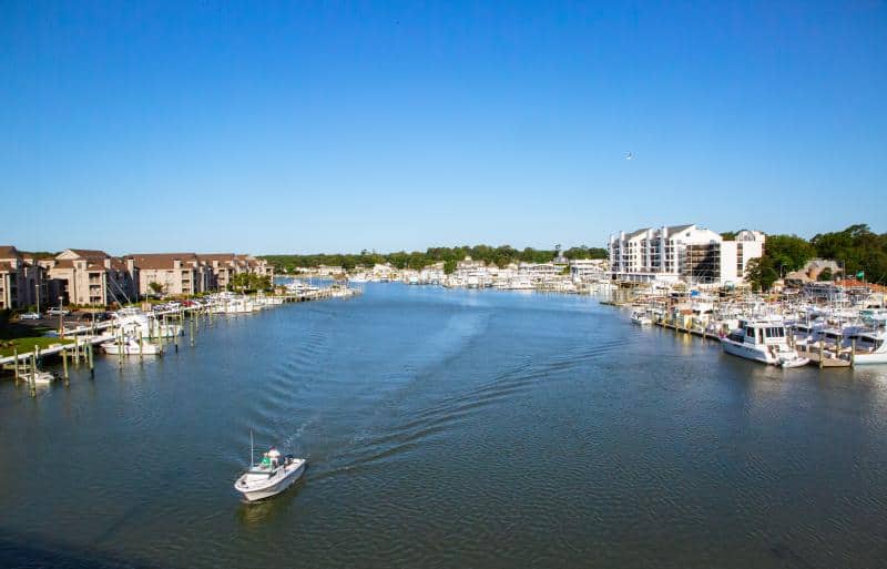 A small boat navigates a wide marina canal lined with docked boats and waterfront buildings under a clear blue sky.