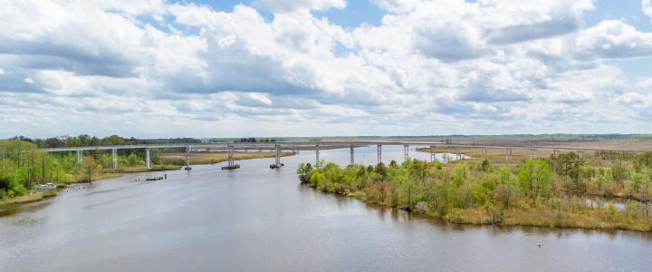 A wide river flows under a long bridge, with a cloudy sky and greenery on both sides.