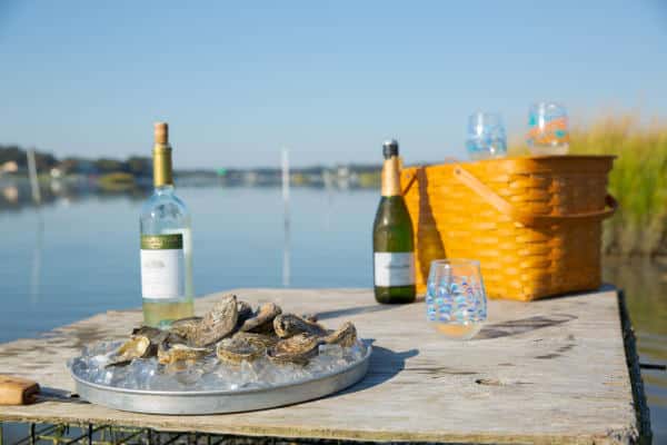 A table by the water holds a tray of oysters, two wine bottles, glasses, and a wicker basket, suggesting an outdoor picnic setting.