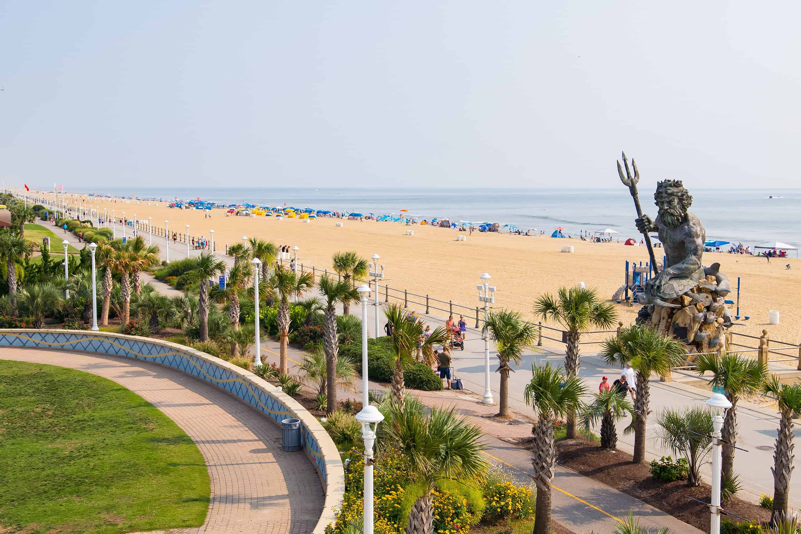 A sandy beach with scattered colorful umbrellas, people relaxing, and a large statue of Poseidon near a promenade lined with palm trees.