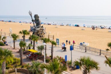 Beach scene with a large King Neptune statue holding a trident, a boardwalk with palm trees, and people walking or lounging on a sandy beach by the ocean.