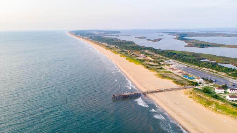 Aerial view of a long sandy beach with a wooden pier extending into the ocean. The shoreline is bordered by grassy areas and inland bodies of water.