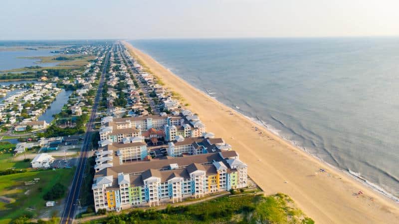 Aerial view of a beachfront residential area with multi-story buildings, a long sandy beach, and calm ocean waves on a clear day.