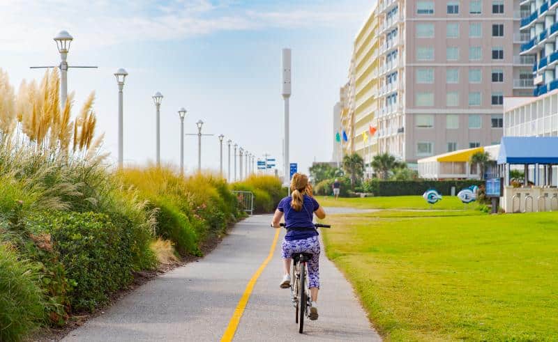 Person riding a bike on a path lined with grass and shrubs, near tall buildings and lampposts.