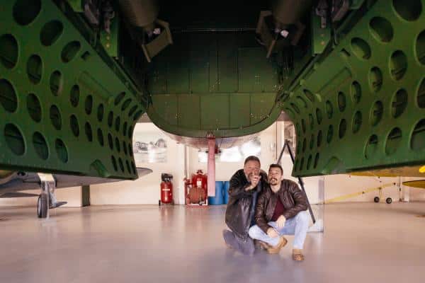 Two people crouch underneath a large green aircraft, pointing at something, in an indoor museum setting.