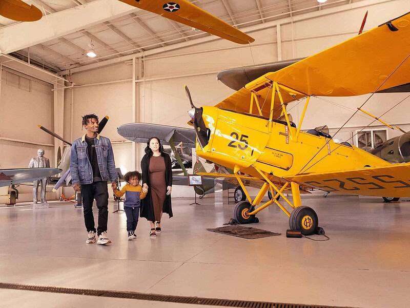 A family strolls through the Military Aviation Museum, featuring vintage airplanes, including a bright yellow bi-plane.
