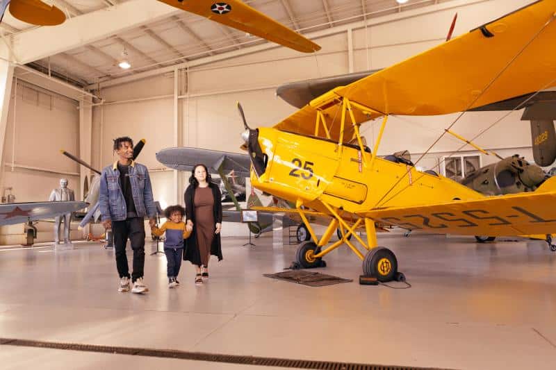 A family of three walks through an aircraft museum hangar, with a yellow biplane and other planes displayed in the background.