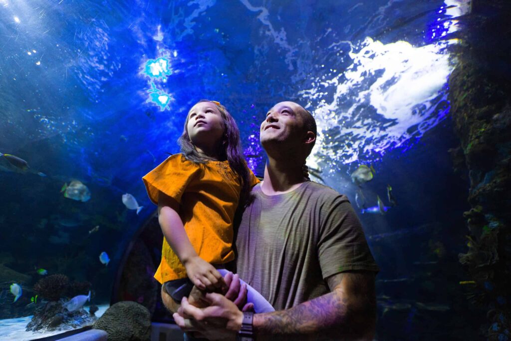 Man holding a child in an aquarium tunnel, both looking upwards with smiles. Blue light and fish surround them, creating an underwater ambiance.