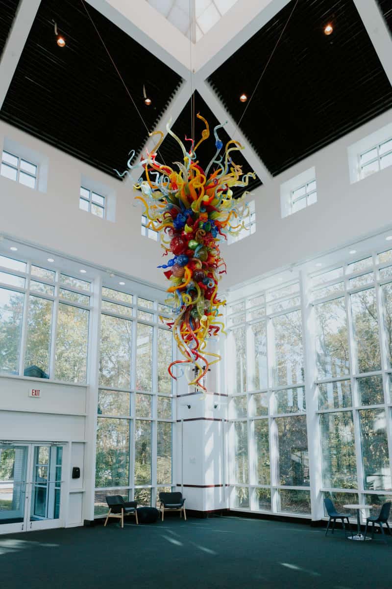 Colorful glass sculpture hanging from the ceiling of a bright, spacious room with large windows and modern seating.