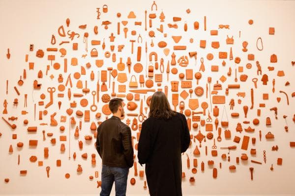 Two people observe a wall display filled with various red objects arranged in an organized pattern.