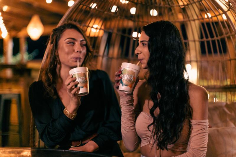 Two people sit in a cozy setting, holding cups and smiling at each other. The background features warm lighting and decorative hanging lights.