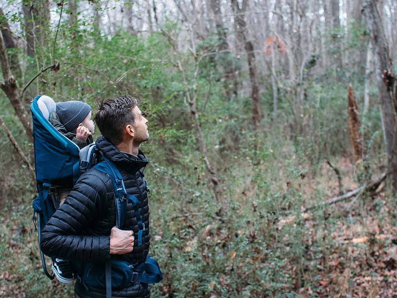 Man hiking in a forest carries a child in a blue backpack carrier. Both wear jackets. Sparse trees and undergrowth surround them.