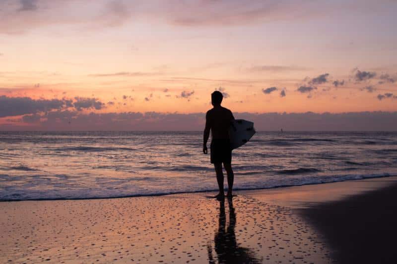 Silhouette of a person holding a surfboard, standing at the shoreline during a colorful sunset.