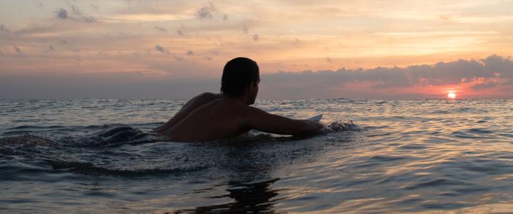 Person lying on a surfboard, facing a sunset over the ocean, with calm waves and cloudy sky in the background.