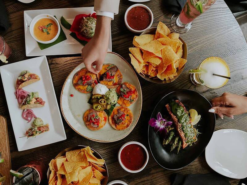 An aerial view of a large spread of Mexican food with hands reaching out, as well as a flight of Margaritas from Side Street Cantina in Virginia Beach.