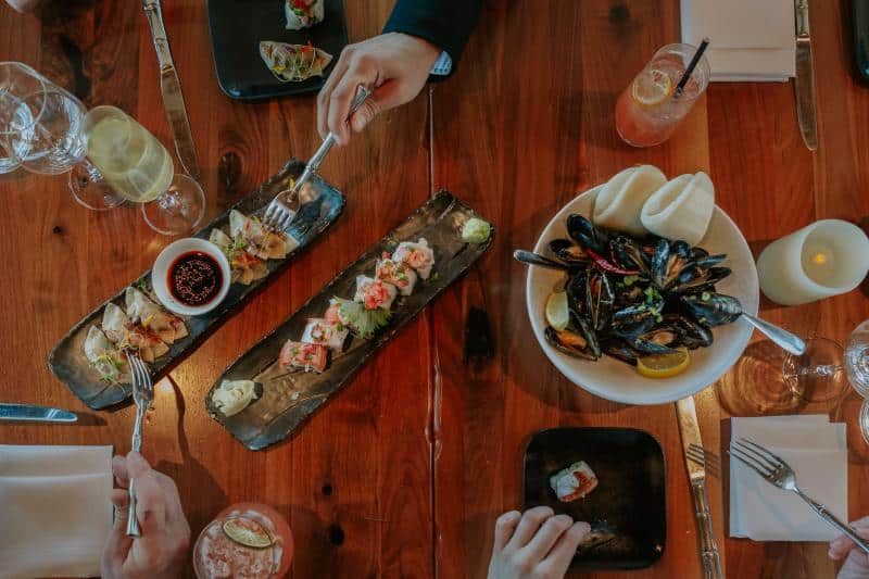 Dining table with assorted dishes including mussels, sushi, and cocktails. Three people are dining, with utensils and glasses visible.
