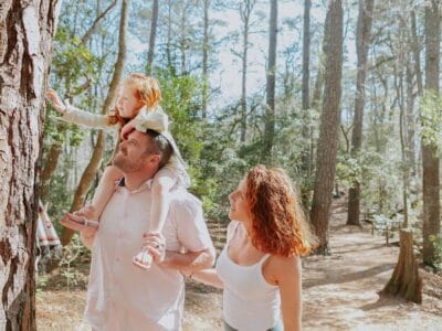 A man carries a young girl on his shoulders as a woman walks beside them. They are in a forest, with trees and sunlight around them. The girl reaches towards a tree trunk.