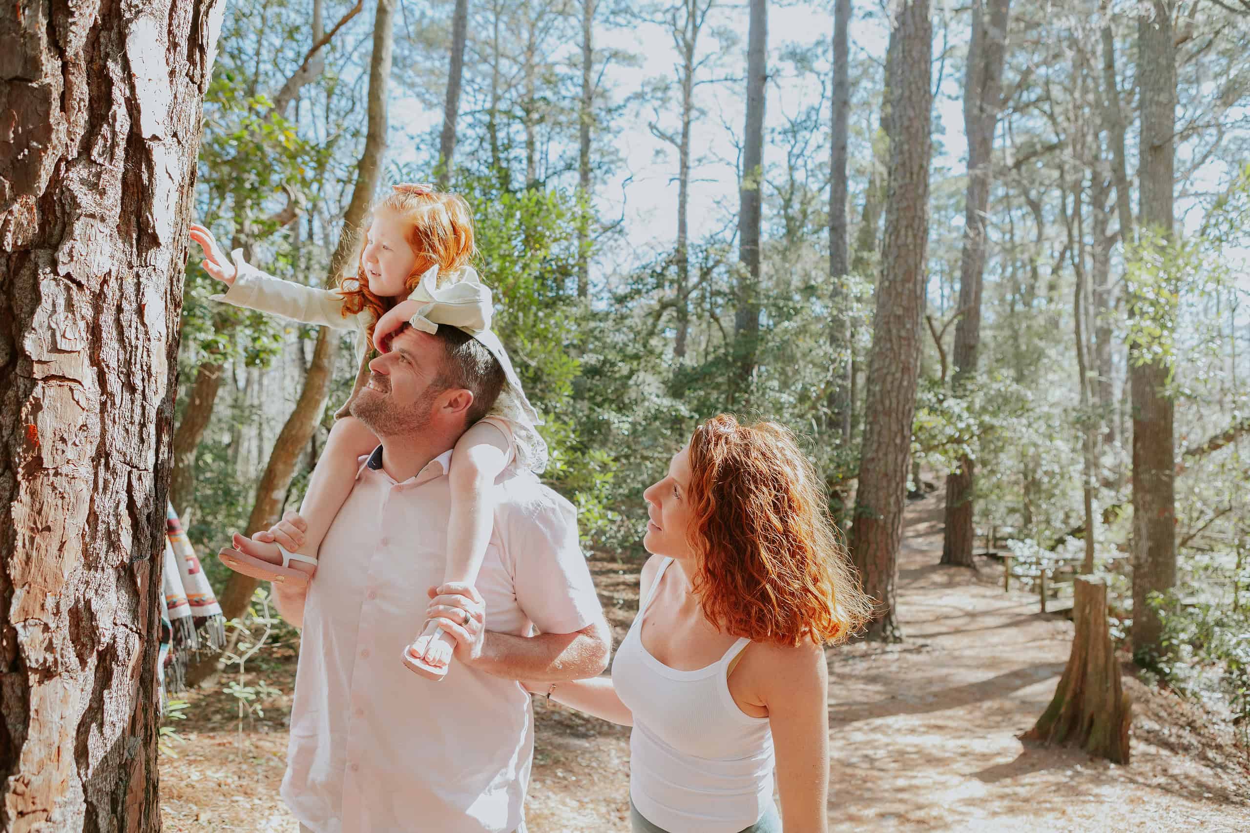 A man carries a young girl on his shoulders as a woman walks beside them. They are in a forest, with trees and sunlight around them. The girl reaches towards a tree trunk.