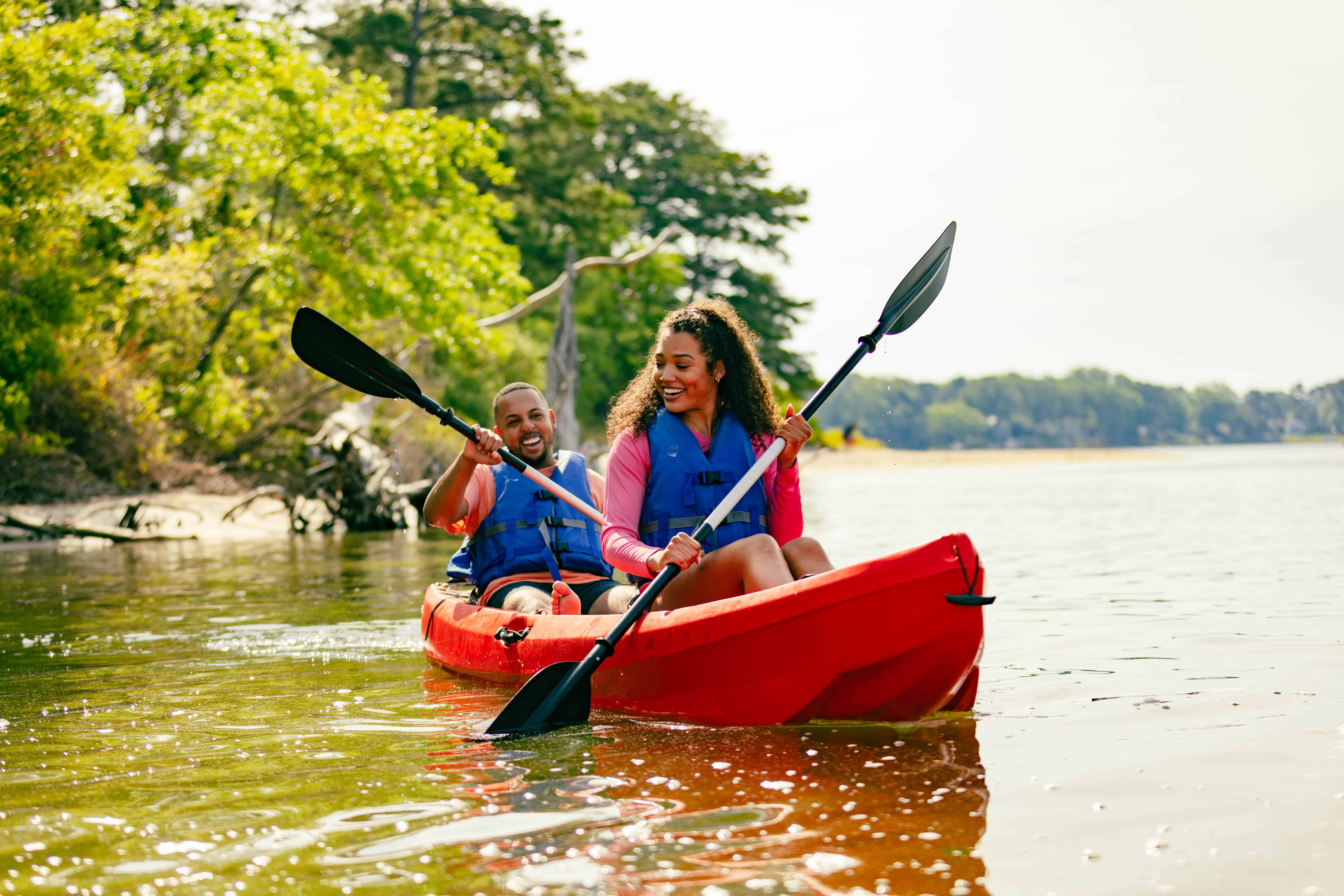 Two people in life jackets kayaking on a calm waterway, surrounded by greenery and trees under a clear sky.