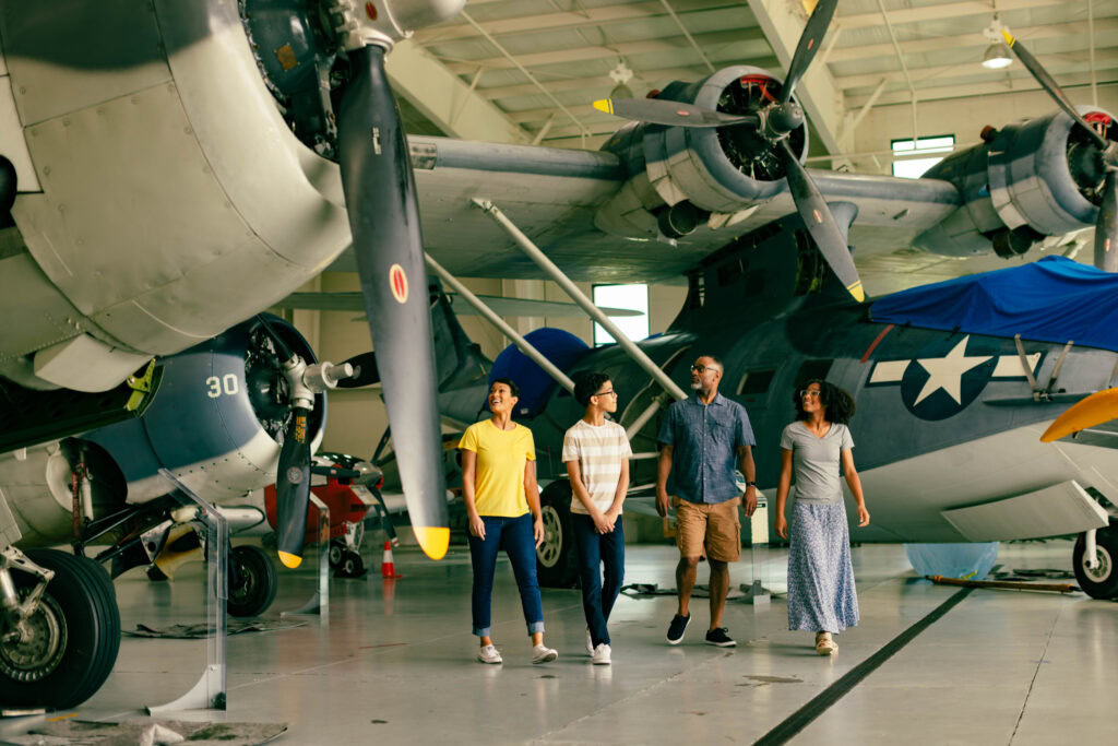 Four people walking in an aircraft hangar, observing vintage planes and propellers under bright lighting.