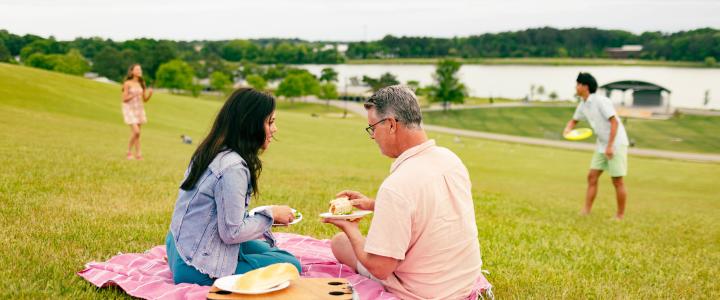 A man and woman sit on a picnic blanket, eating. Another man and woman play catch with a frisbee in the background. A lake and trees are visible in the distance.