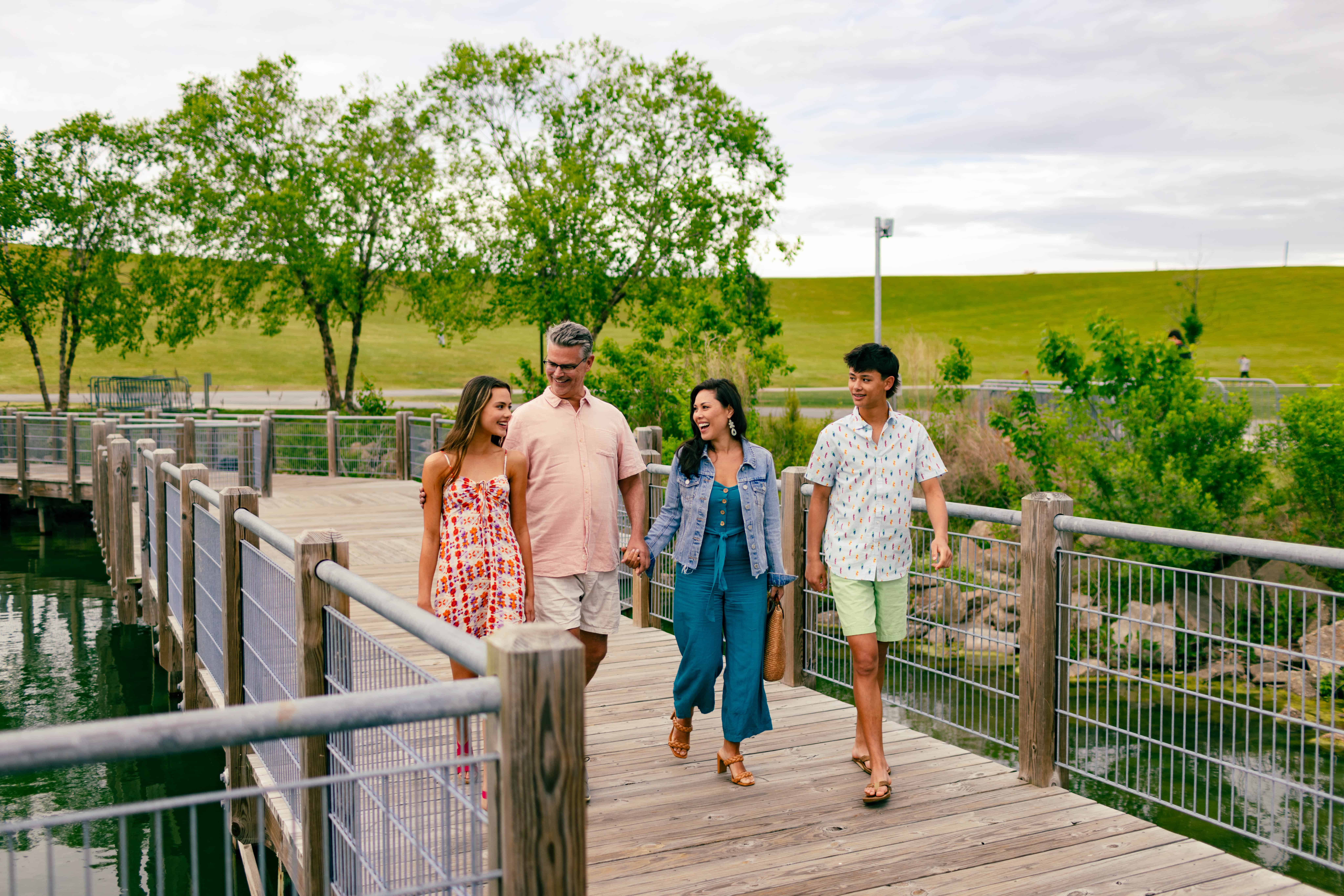 Four people walking and talking on a wooden bridge in a park, surrounded by trees and grass.