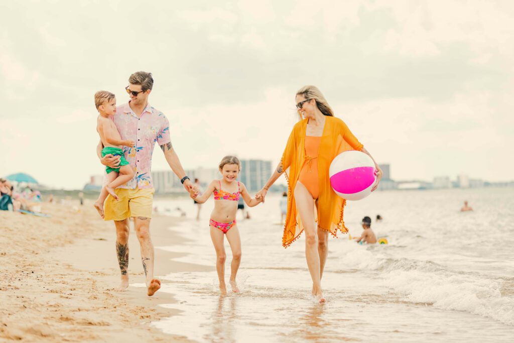 Family of four walks on the beach, holding hands. The mother carries a beach ball, and the father carries a toddler. A young girl walks between them. The sea and people are visible in the background.