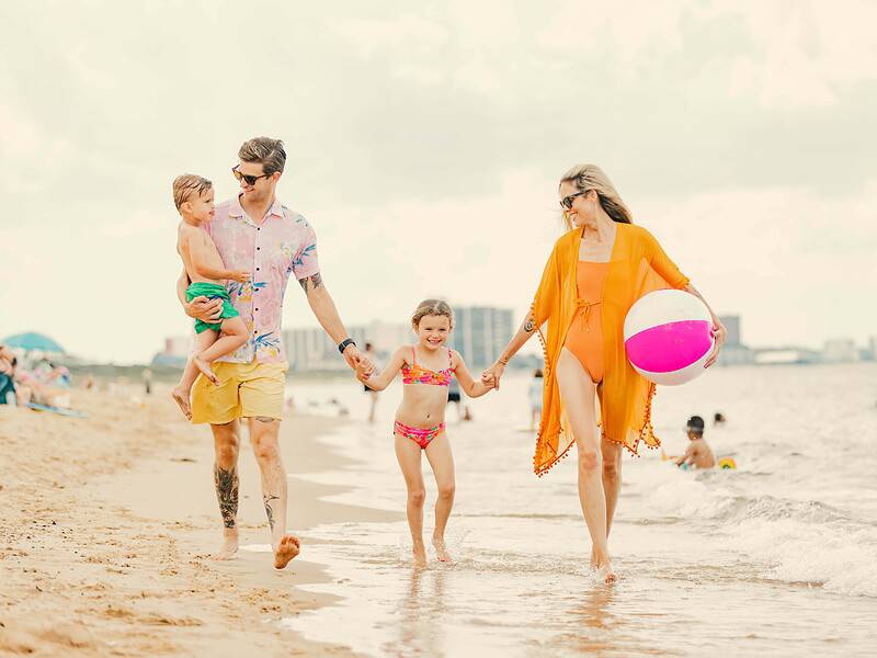 Family of four walks on the beach, holding hands. The mother carries a beach ball, and the father carries a toddler. A young girl walks between them. The sea and people are visible in the background.