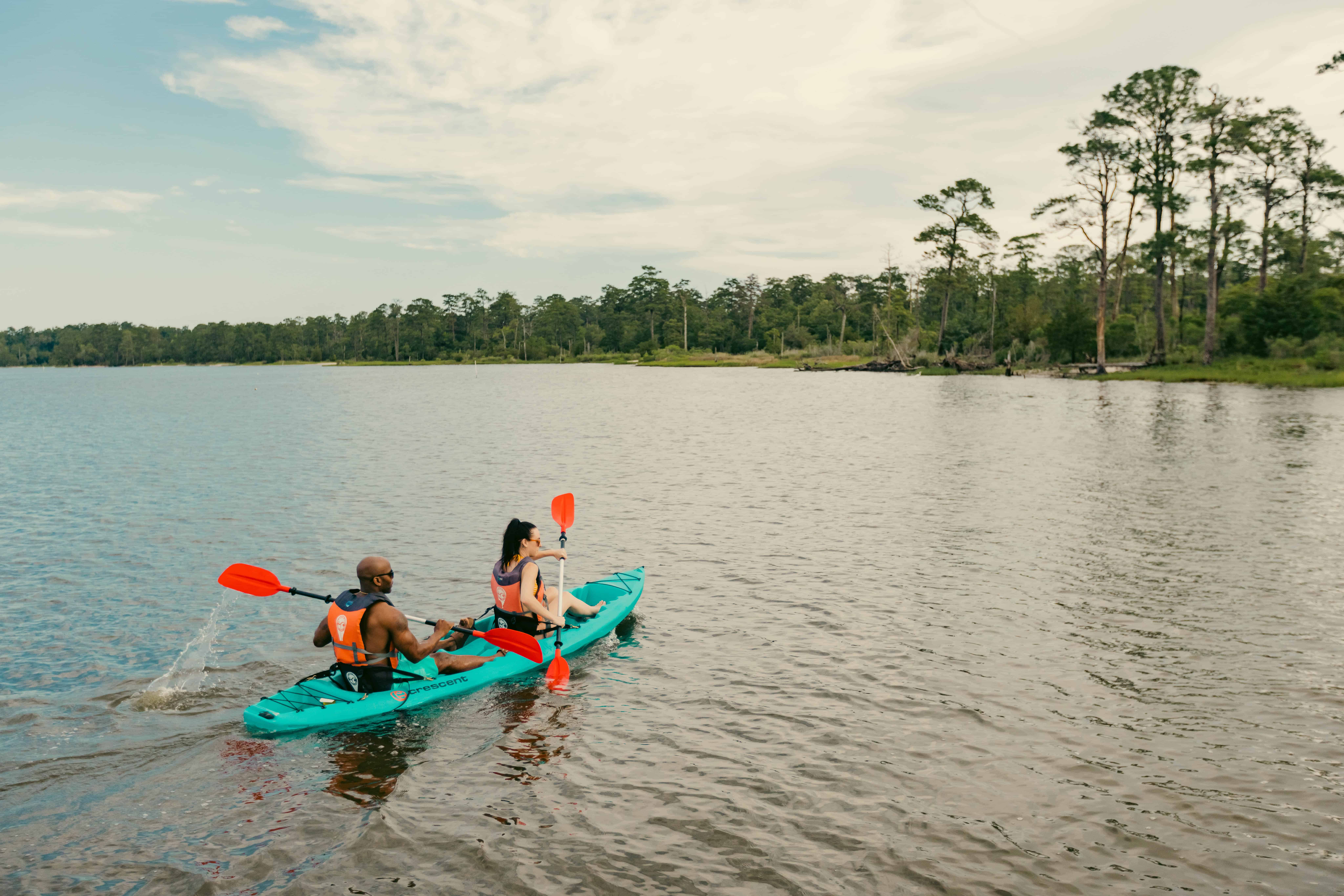 Two people kayaking on a calm lake near a forested shore under a partly cloudy sky. Both wear life vests and use orange paddles.