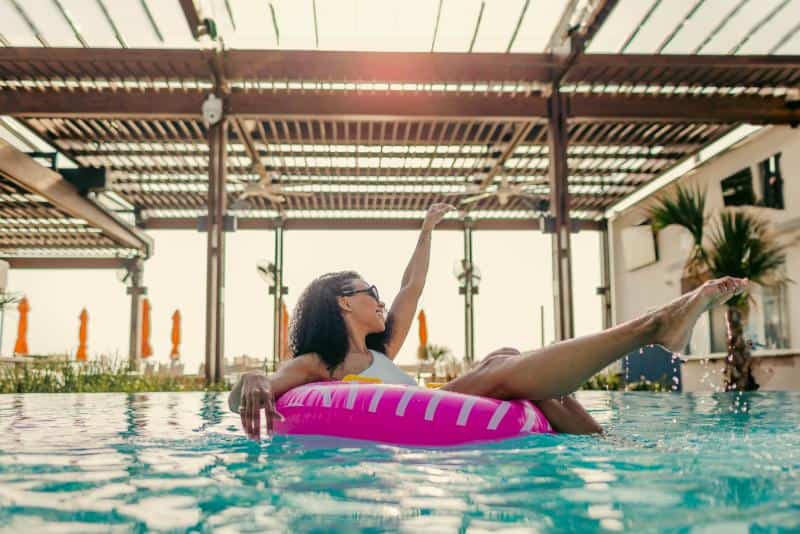Person lounging on a pink inflatable pool float, wearing sunglasses, under a pergola with palm trees nearby.