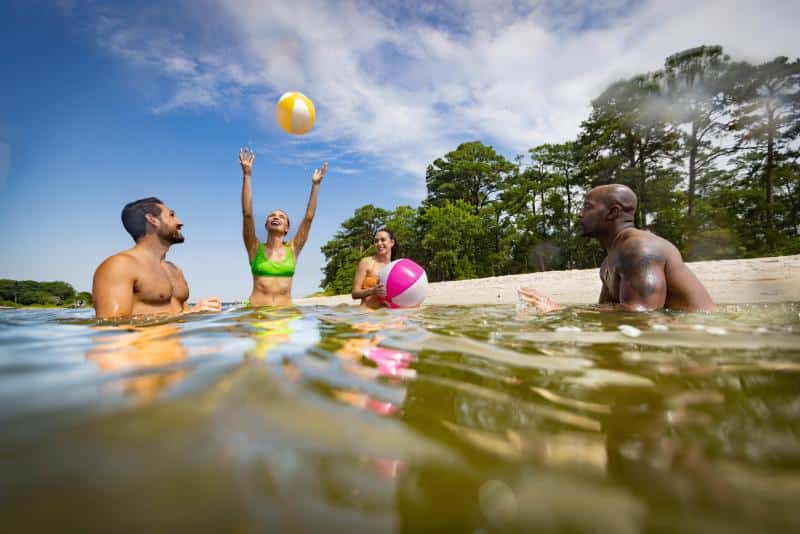 Four people in the water, playing with beach balls near a sandy shore; trees visible in the background under a blue sky with clouds.