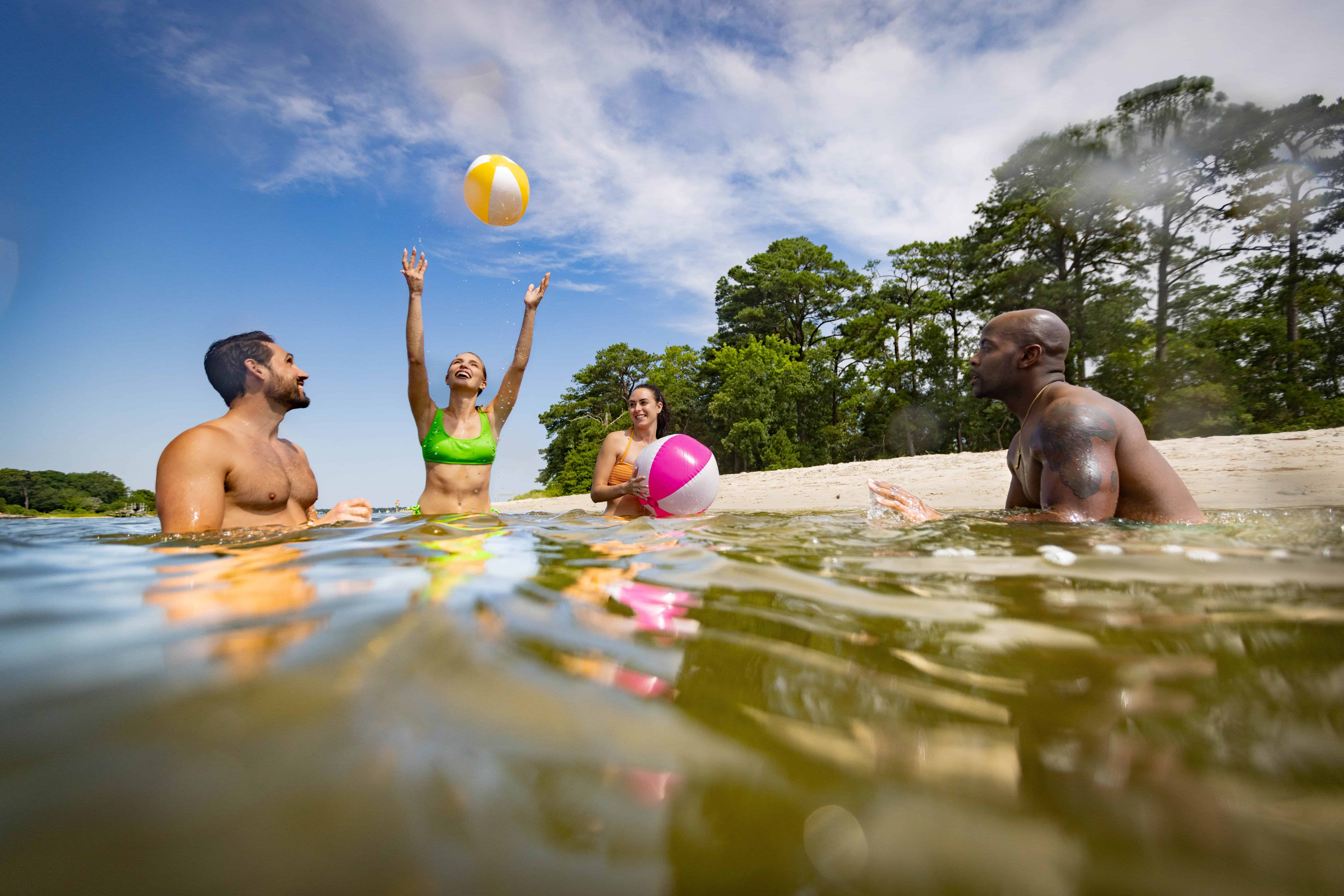 Four people are enjoying a game with colorful beach balls in a sunny, shallow lake near a sandy shore with trees.