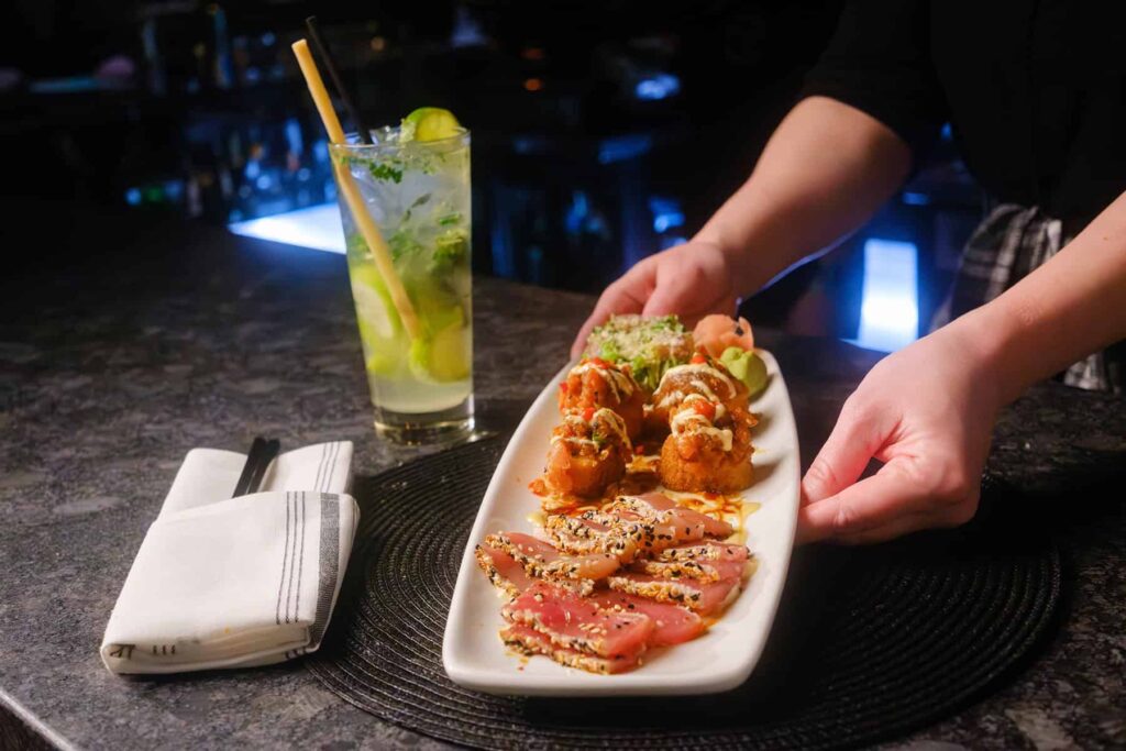 Person serving a platter of sesame-crusted tuna slices and fried shrimp on a round placemat. A beverage with mint and lime is next to a napkin on a dark countertop.