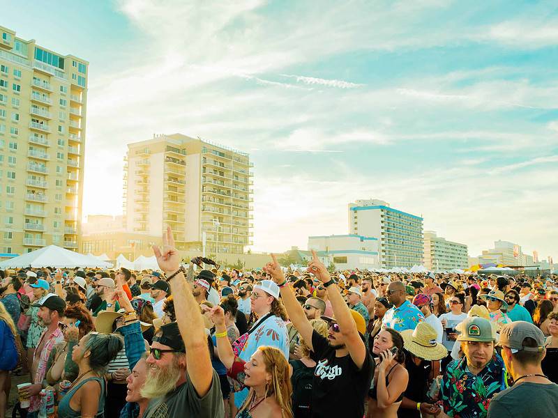 A large crowd of people gathered outdoors near tall buildings, enjoying a sunny day, with some raising their arms in the air.