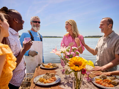 A group of people stand around a table with seafood by a waterfront. They are smiling and engaged in conversation under a clear blue sky. Flowers decorate the table.