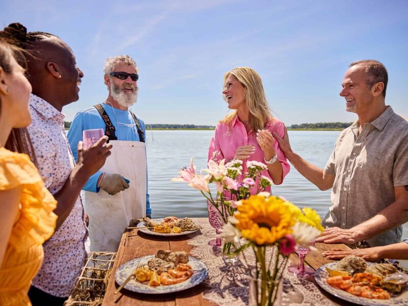 A group of people stand around a table with seafood by a waterfront. They are smiling and engaged in conversation under a clear blue sky. Flowers decorate the table.